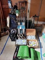 Full view of golf clubs in bag, golf shoes, three boxes of golf balls, and putting green mat.
