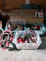Collection of Christmas decorations including two metal tree stands, three plastic bins filled with ornaments, garland, lights, and a large box housing the artificial tree.