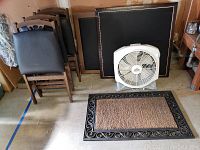 Image showing four wooden folding chairs with padded black vinyl seats, two vintage wooden bridge card tables with black leather-lined surfaces leaning against the wall, white box fan in front, and entry rug placed on floor.