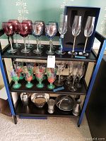 Full view of all items on black shelving including cranberry glasses, blue glasses, champagne flutes, frog glasses, martini shaker, and pewter trays.