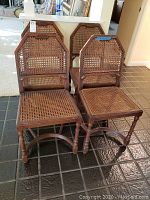 Four vintage wooden chairs with cane woven backs and seats, arranged in two pairs on a tiled floor indoors under natural light.