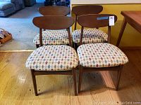 Four mid century wooden dining chairs with curved backrests, pattered upholstery on seat cushions, arranged on wooden floor.