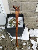 Walking stick positioned vertically next to a planter outside with patchy snow, showing the natural wood texture and the top handle shape.