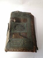 Photo of the front cover showing embossed green leather with decorative oval scene and wear including tears and chips.