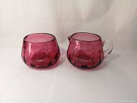 Front view of cranberry art glass creamer jug and sugar bowl set against a neutral background.