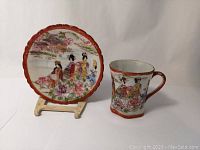 Photo of demitasse cup with hand-decorated Japanese geisha pattern next to matching saucer, both with reddish-orange scalloped rims.