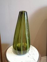 Front angle view of the green art glass vase placed on a white marble surface, showing its tapered shape and vertical swirl pattern.