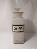 Front view of white apothecary bottle with glass stopper and bordered label reading 'POT. CARB.'