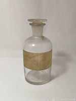 Front view of the clear glass apothecary bottle with paper label and flat round glass stopper on top, standing upright on a plain background.