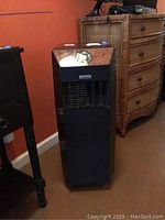 Full view of black Amcor portable air conditioner next to furniture against an orange wall.