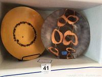 Two round designer plates on stands shown side by side in a display shelf. One plate is yellow with a circular pattern of black dots, the other is dark gray with abstract tan and black shapes, each approximately 14 inches in diameter.