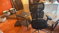 Ergonomic office chair with mesh back and headrest, black metal side table with two shelves, wire basket, and adjustable height paper stand visible