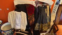Photo showing the collection of vintage evening garments including the black Colombian shawl with long macramé fringe, the beaded white jacket, and skirts hanging together on hangers.
