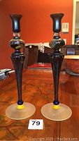 Pair of tall signed glass candle holders with textured black stems, frosted broad bases with blue and yellow glass accents, and rounded multicolored decorative midsections
