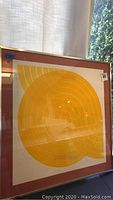 Framed abstract art print with concentric yellow shapes and reddish frame.