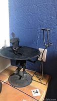 Side view of black metal art sculpture with two human figures and platform, placed on tiled floor against blue wall.