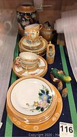 Photo showing a stacked set of luster ware dinner plates, cups, saucers, and additional ceramic items including bird and house figurines arranged on a table.