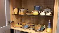 Full cabinet shelf showing various luster ware pottery pieces including teapots, pitchers, small bowls, and salt/pepper shakers.