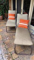 Pair of beige fabric outdoor chaise lounge chairs with orange pillows placed on stone patio outside a building.