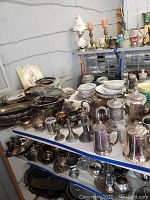 Overall view of shelves loaded with various silver plated platters, footed bowls, cups, coffee pots, and additional serving items in a warehouse setting