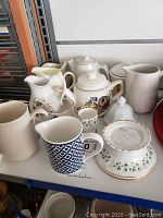 Variety of ceramic creamers, pitchers, tea pots, and vases shown on a shelf.