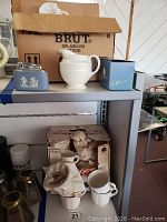 Photo showing blue Wedgwood creamer, lidded sugar bowl, trinket box, and metal lighter placed on a shelf with boxes behind them.