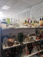 Wide view of shelves showing various glassware, ceramics, oil lamps, and decorative items including egg coddlers, pitchers, and candle holders.