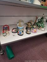 Photo of nine assorted snow globes displayed on a white shelf with various themes and figures.