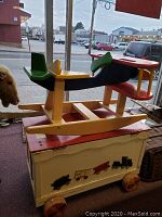 Side view of wooden airplane rocker on top of white toy box with colorful wheels and lid design.