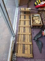 Full view of the antique wooden toboggan showing its length and construction with multiple slats laid lengthwise and crosswise, placed beside other items in the room.