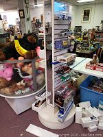 Front side of the white swivel display rack showing slatwall panels with hooks and shelves holding various boxed items, bear plush, and other small objects, demonstrating its use before clearing.