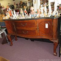 Front view of the marble top buffet showing serpentine front, curved cabinet doors, two drawers, and Chippendale style wooden legs and carvings. Various figurine items on top not included.