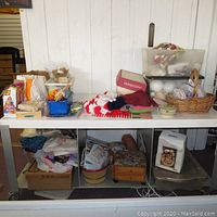 Wide view of crafting materials on white table and shelves including yarn bundles, plastic containers with foam balls, and boxes with crafting kits.