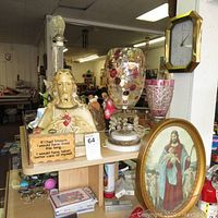 Wide view showing the bust of Jesus, decorative glass vase, pink goblet, framed religious art, and clock in a store setting.