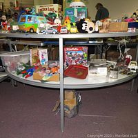 Wide view of vintage toys on two-tiered shelf including plastic figurines, die-cast cars, plush toy, dolls, boxes and a green roof plastic house.