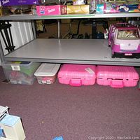 View underneath a table showing two pink cases, a bin, and a Barbie camper vehicle on a shelf.