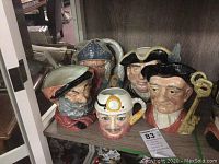 Four Toby jugs arranged on a wooden shelf, showing front faces including one with black hat, one with white hat and monocle, one with a tricorn hat, and one with a blue patterned cap.