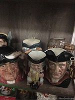 Shelf display of five Royal Doulton figural toby jugs and mugs. Left to right front includes Night Watchman jug, Jolly Toby mug, Romeo mug, with additional mugs behind.