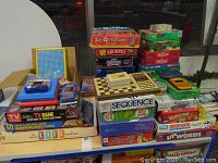 Photo shows a large assortment of board games and puzzles stacked on a table including Life, Sequence, Yahtzee, Monopoly Jr, Upwards, 20 Questions, Trumpet and various puzzle boxes.
