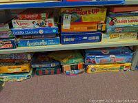 Photo of assorted games including Scrabble, Chinese Checkers, Goosebumps, Puzzle Roll Up mat, Beginners Bridge, Clue, Carmen Sandiego, and several other boxed games stacked on a shelf.