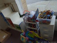 Wide shot of white dollhouse-style organizer with colored drawers, plastic bins stacked with toys, bins filled with dinosaur and animal figurines, and toy track sets.
