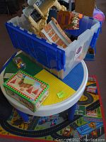 Photo of children’s Lego play table with multi-color base plates (blue, yellow, green) featuring multiple toys and boxes on top including Lincoln Logs in original box and plastic toys.