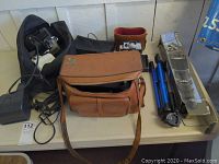 Photo showing two Polaroid cameras, tan leather camera bag, blue tripod, and black charger-type item on table.