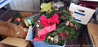 Blue plastic bin filled with artificial Christmas wreaths decorated with pinecones, red berries, wooden balls, and large red velvet bow, surrounded by cardboard boxes with various holiday items including wrapping paper and ornaments.