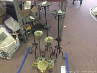Seven metal candle holders arranged on floor, showing varying heights and designs including tall black holders, silver-toned holders, and a gold-toned piece