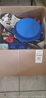 Photo showing open cardboard box filled with blue silicone baking pans, various cooking utensils including spatulas and brushes with red and natural wood handles, and packaging boxes.