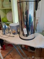 Ironing board with iron, small Minions wastebasket, and stainless steel trash can on top of ironing board.