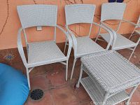 Three white wicker chairs and one matching side table arranged outside on tile floor near wall.