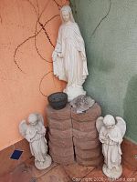 Full view: Madonna statue atop stacked paver bricks flanked by two angel statues with decorative bowl and cherub plaque visible