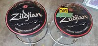 Two chrome metal barstools with round black cushioned seats featuring the Zildjian logo and text 'Genuine Turkish Cymbals'. The seats have decorative red borders and floral emblem.
